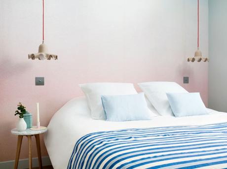 mur tie and dye rose dans une chambre à coucher. Lit blanc avec un plaid rayé bleu et blanc, deux suspension cartonnées de part et d'autre du lit. Un tabouret bois et marbre en guise de chevet.