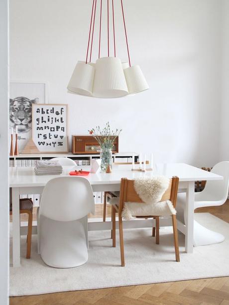 Une salle à manger conviviale, chaises dépareillés, blanches et bois, des bibliothèques basses près de la table, accumulation de suspensions blanches fils rouges