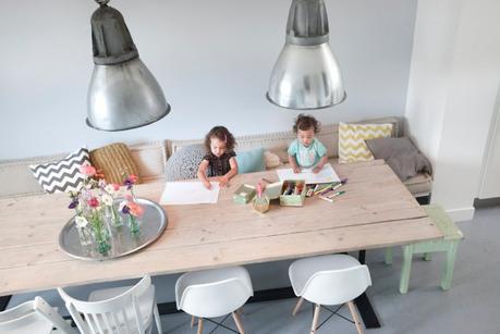 grande table de repas en bois, deux chaises RAR eames blanches, deux grandes suspensions industrielles, une banquette avec des coussins graphique et deux jeunes enfants qui dessinent